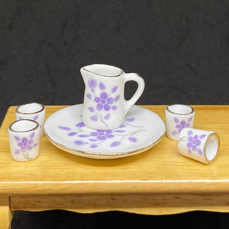 May include: A miniature tea set with a white pitcher and four cups, all decorated with a purple floral pattern and gold trim. The set is on a wooden table.