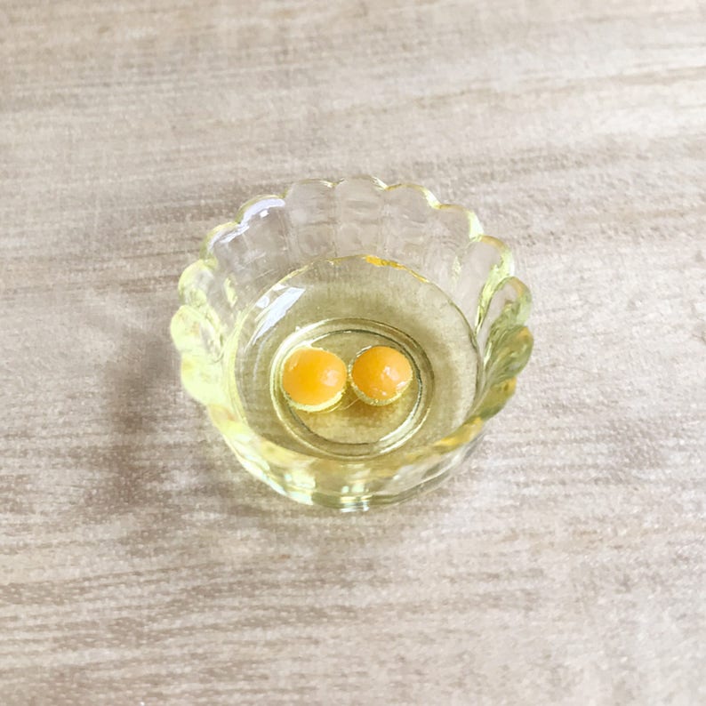 Op de afbeelding: Een kleine, heldere glazen kom met een geschulpte rand, met een gele vloeistof en twee ronde, oranje-gele objecten. De kom staat op een lichtgekleurd, getextureerd oppervlak.