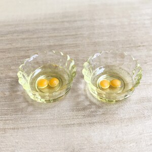 Op de afbeelding: Twee kleine, heldere glazen kommen met geschulpte randen, elk met twee kleine, ronde, gele objecten. De kommen staan op een lichtgekleurd, getextureerd oppervlak. De kommen hebben een lichte gele tint.