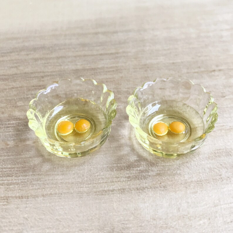 Op de afbeelding: Twee kleine, heldere glazen kommen met geschulpte randen, elk met twee kleine, ronde, gele objecten. De kommen staan op een lichtgekleurd, getextureerd oppervlak. De kommen hebben een lichte gele tint.