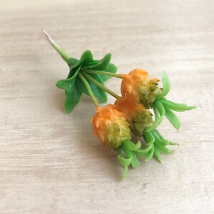 May include: A close-up of a small, artificial flower arrangement. The arrangement features three orange and green flower buds with green leaves. The stem is a light green color.