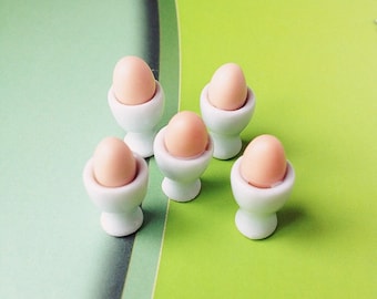 May include: Five miniature white ceramic egg cups with pink eggs inside. The egg cups are arranged in a semi-circle on a green surface.