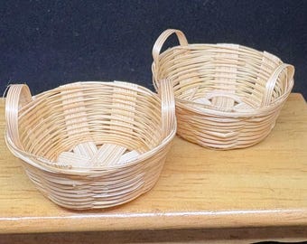 Op de afbeelding: Twee kleine, ronde, geweven manden met handvatten. De manden zijn gemaakt van lichtgekleurd, natuurlijk ogend materiaal, mogelijk bamboe of riet. Ze staan op een lichtbruin oppervlak tegen een donkere achtergrond. De manden hebben een eenvoudig, rustiek ontwerp.