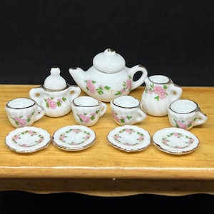 May include: A miniature white porcelain tea set with pink floral designs. The set includes a teapot, creamer, sugar bowl, four teacups, and four saucers. Each piece has gold trim, creating a delicate and charming aesthetic.