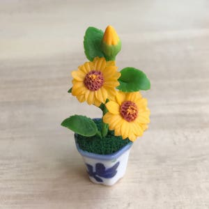 以下が含まれることがあります： 青い花の模様のミニチュアセラミックポットには、小さなヒマワリの植物が入っています。植物には、茶色の中心部、緑の葉、閉じたつぼみのある2つの開いた黄色のヒマワリがあります。ポットは緑色の苔で満たされています。