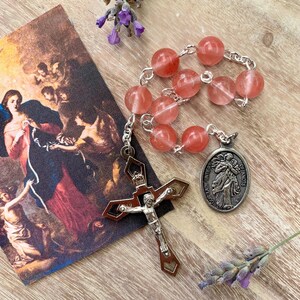 May include: A pink and white rosary with a silver crucifix and a silver medal of the Virgin Mary, Untier of Knots. The medal has the text "Our Lady Untier of Knots".