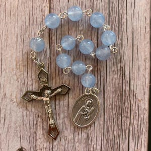 May include: A rosary with light blue beads and a silver crucifix. The crucifix features a detailed depiction of Jesus. A silver medal with the image of St. Lucy is also attached.