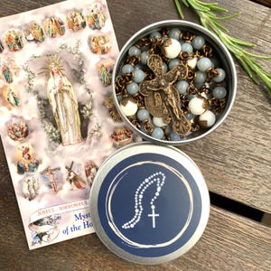 May include: A rosary with blue and white beads and a bronze crucifix is displayed in a small tin. A card with religious imagery and the words "Joyful - Sorrowful" is next to the tin. The tin has a blue lid with a white rosary design.