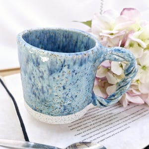 May include: A blue speckled ceramic mug with a twisted handle, resting on a notebook with a silver spoon. The mug has a white base. A bouquet of pink and white flowers is in the background. The mug is suitable for hot drinks.
