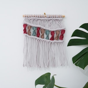 May include: A macramé wall hanging with a light grey background and a wooden dowel. The design includes red, teal, and brown twisted rope accents. The bottom features long, dangling fringe. This handcrafted piece adds a touch of bohemian style to any room.