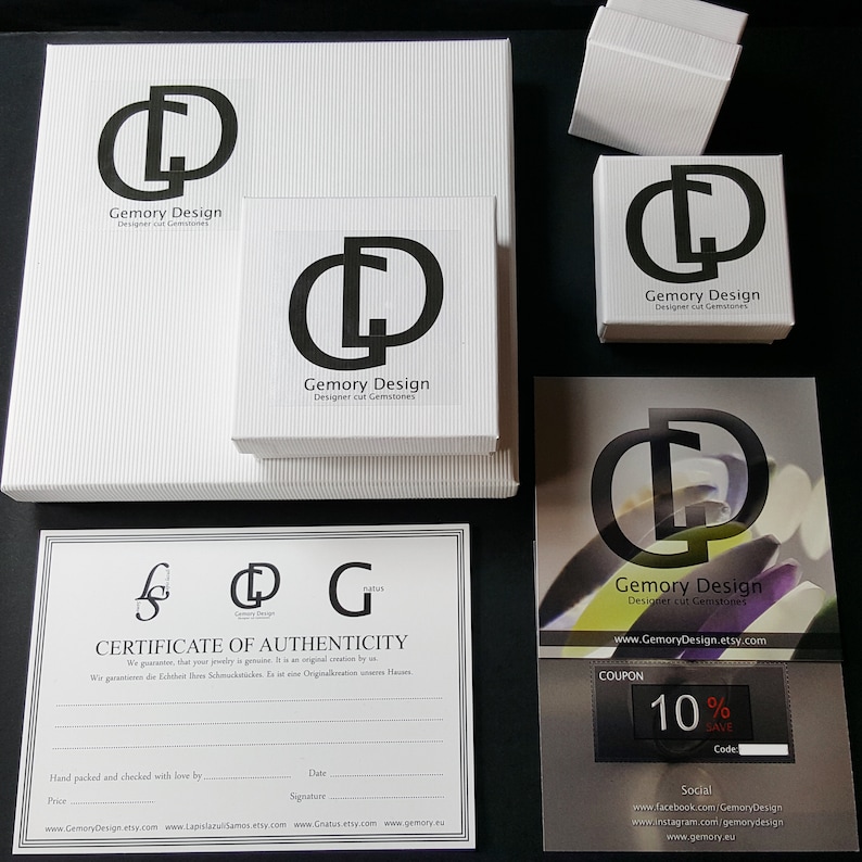 Op de afbeelding: Een witte doos met het logo "Gemory Design" en de tekst "Designer cut Gemstones". De doos staat op een wit oppervlak. Er is een certificaat van echtheid met de tekst "Gemory Design" en "Certificate of Authenticity". Er is ook een coupon met de tekst "Gemory Design" en "10% SAVE".