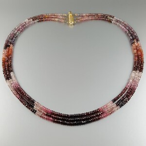 May include: A multi-strand necklace made of pink, red, and purple gemstone beads. The necklace is made of three strands of beads, each strand is a different colour. The beads are small and round.