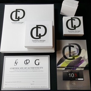 May include: White boxes and packaging with the "Gemory Design" logo, including a certificate of authenticity and a coupon for 10% off. The logo is a stylized "GD" in black. The packaging is on a black surface.