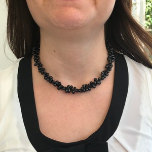 May include: A black beaded necklace with a unique, clustered design. The necklace is worn against a white blouse with black trim. The beads have a glossy finish, creating a striking contrast against the wearer's skin and clothing.