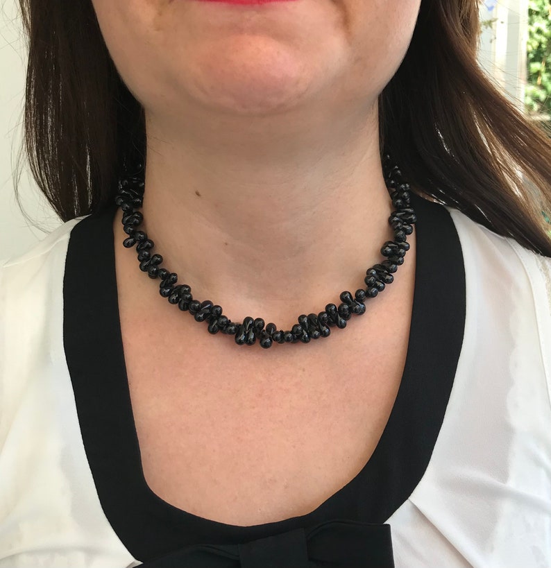 May include: A black beaded necklace with a unique, clustered design. The necklace is worn against a white blouse with black trim. The beads have a glossy finish, creating a striking contrast against the wearer's skin and clothing.