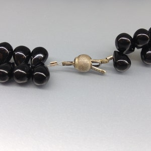 May include: Close-up of a black beaded necklace with a silver clasp. The beads are teardrop-shaped and have a glossy finish. The clasp is a simple, functional design.