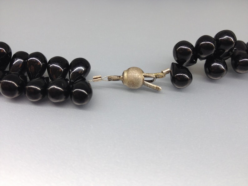 May include: Close-up of a black beaded necklace with a silver clasp. The beads are teardrop-shaped and have a glossy finish. The clasp is a simple, functional design.