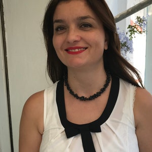 May include: A woman wearing a white sleeveless top with a black bow and trim. She is also wearing a black beaded necklace. The woman has brown hair and is smiling, with red lipstick.
