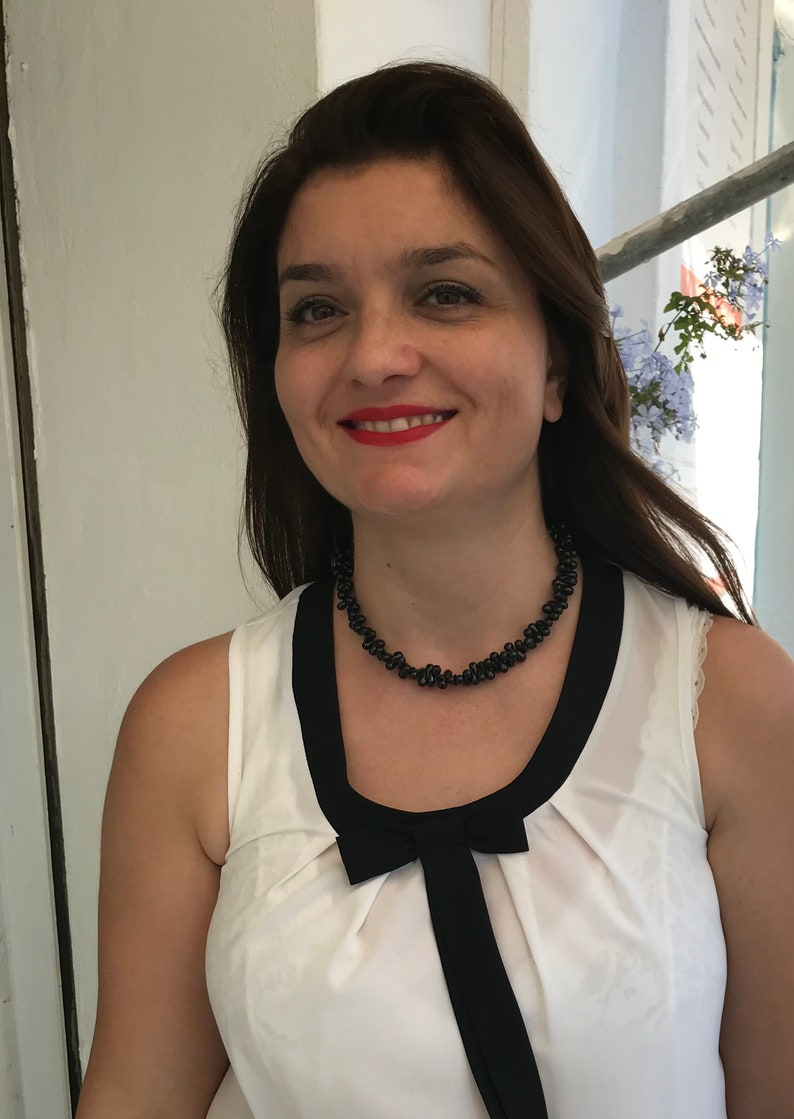 May include: A woman wearing a white sleeveless top with a black bow and trim. She is also wearing a black beaded necklace. The woman has brown hair and is smiling, with red lipstick.