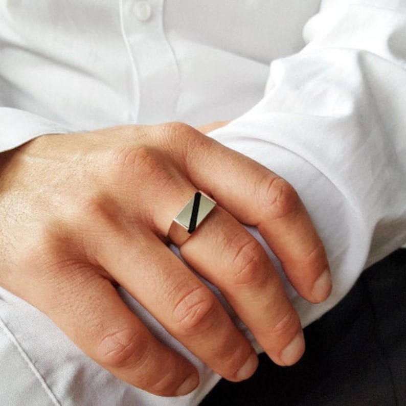 Op de afbeelding: Een zilveren ring met een zwarte rechthoekige inleg. De ring wordt gedragen aan de wijsvinger van een hand.