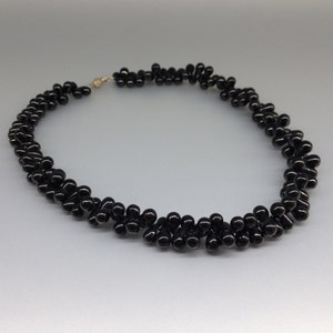 May include: A black beaded necklace with a gold-colored clasp. The necklace is made of small, glossy, teardrop-shaped beads arranged in a twisted pattern. The beads are tightly clustered, creating a textured appearance. The necklace is displayed on a light gray background.