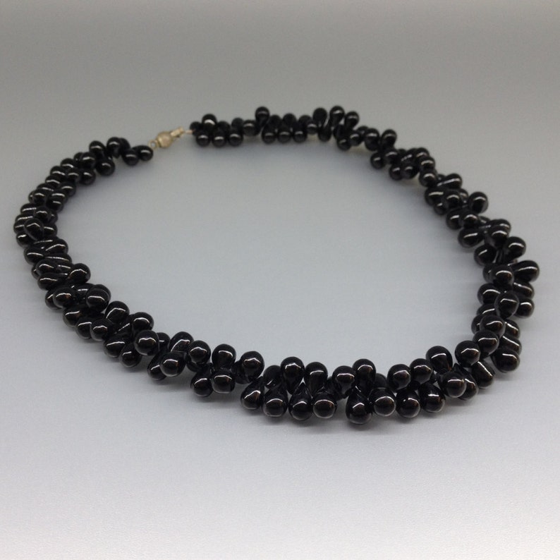 May include: A black beaded necklace with a gold-colored clasp. The necklace is made of small, glossy, teardrop-shaped beads arranged in a twisted pattern. The beads are tightly clustered, creating a textured appearance. The necklace is displayed on a light gray background.