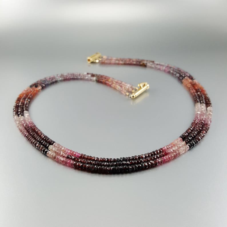 May include: A multi-strand necklace made of pink, red, and purple gemstone beads. The necklace is made of three strands of beads, each strand is a different colour. The necklace is made of small, round beads.