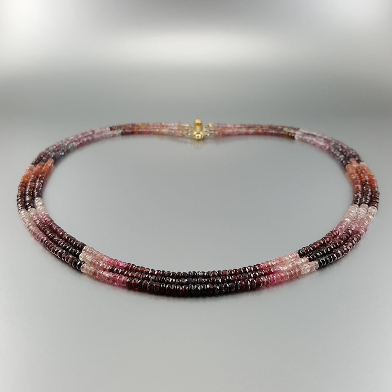May include: A multi-strand necklace made of pink, red, and purple gemstone beads. The necklace is made of small, round beads and has a gold clasp.