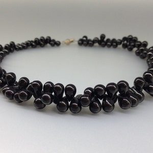 May include: A black beaded necklace with a teardrop design. The necklace features multiple strands of glossy, dark beads, creating a layered effect. The clasp is gold-toned, and the necklace is displayed against a light background.