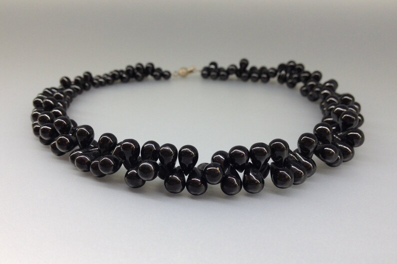 May include: A black beaded necklace with a teardrop design. The necklace features multiple strands of glossy, dark beads, creating a layered effect. The clasp is gold-toned, and the necklace is displayed against a light background.