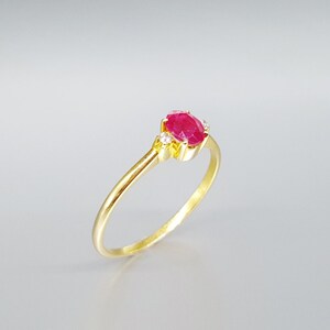 Könnte beinhalten: Ein goldener Ring mit einem roten ovalen Rubin-Edelstein und zwei kleineren weißen Edelsteinen auf der Ringschiene.