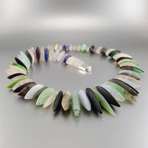 May include: A necklace made of various colored stones, each shaped like a teardrop. The stones are strung together on a silver chain. The necklace is laid out in a semi-circle on a gray surface.