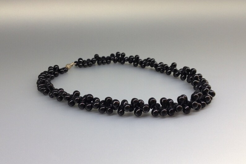 May include: A black beaded necklace with a silver clasp. The necklace is made of small, round, glossy black beads arranged in a clustered pattern. The beads are strung together to form a curved shape.
