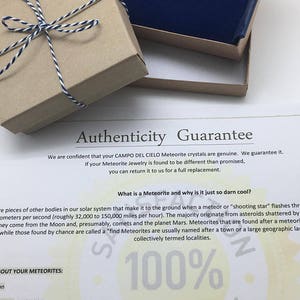 May include: A close-up of a brown cardboard box with a blue velvet box inside. The boxes are tied with a white and black twine. The text on the white paper reads "Authenticity Guarantee" and "What is a Meteorite and why is it just so darn cool?" The text also includes "100% Guarantee".