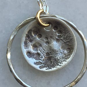 K&ouml;nnte beinhalten: Eine silberne Anh&auml;nger Halskette. Sie zeigt einen strukturierten, kreisf&ouml;rmigen Anh&auml;nger mit geh&auml;mmertem Design und kleinen L&ouml;chern, der in einem gr&ouml;&szlig;eren, geh&auml;mmerten Silberring h&auml;ngt. Ein goldfarbener B&uuml;gel verbindet den Anh&auml;nger mit einer Silberkette.