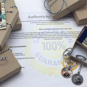 May include: A close-up of a small, brown box tied with a white and black ribbon. The box is labeled "About Your Meteorites" and contains a small piece of meteorite. The image also shows a piece of meteorite jewelry, a blue velvet pouch, and a paper with the text "100% Guarantee" and "Authenticity Guarantee".