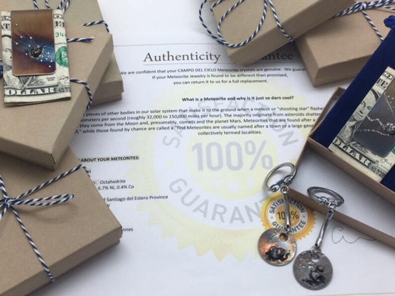 May include: A close-up of a small, brown box tied with a white and black ribbon. The box is labeled "About Your Meteorites" and contains a small piece of meteorite. The image also shows a piece of meteorite jewelry, a blue velvet pouch, and a paper with the text "100% Guarantee" and "Authenticity Guarantee".