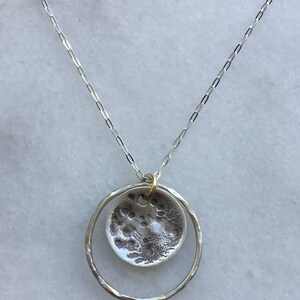 K&ouml;nnte beinhalten: Eine silberne Halskette mit einem Anh&auml;nger mit einem strukturierten, kreisf&ouml;rmigen Design, das an den Mond erinnert, der in einem geh&auml;mmerten Silberring h&auml;ngt. Die Halskette hat eine feine Kette und einen goldfarbenen Verbinder.