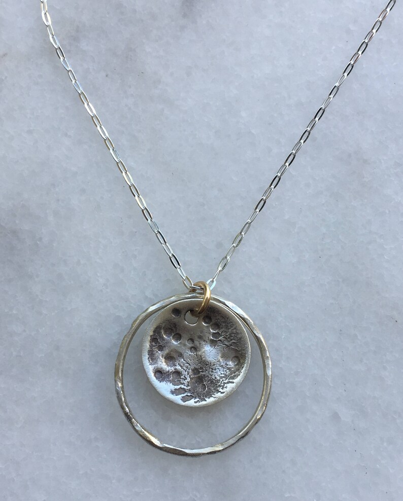 K&ouml;nnte beinhalten: Eine silberne Halskette mit einem Anh&auml;nger mit einem strukturierten, kreisf&ouml;rmigen Design, das an den Mond erinnert, der in einem geh&auml;mmerten Silberring h&auml;ngt. Die Halskette hat eine feine Kette und einen goldfarbenen Verbinder.
