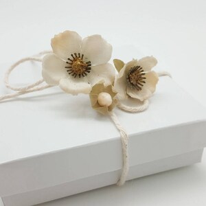 May include: A white gift box with a white string bow and three white paper flowers with gold centers.