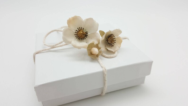 May include: A white gift box with a white string bow and three white paper flowers with gold centers.