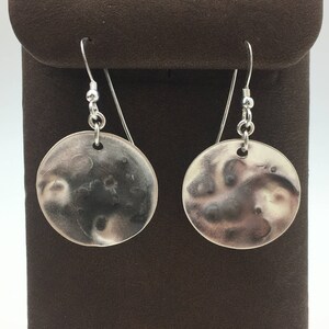 May include: A pair of silver dangle earrings with a textured, moon-shaped design. The earrings are hanging from a brown velvet background.