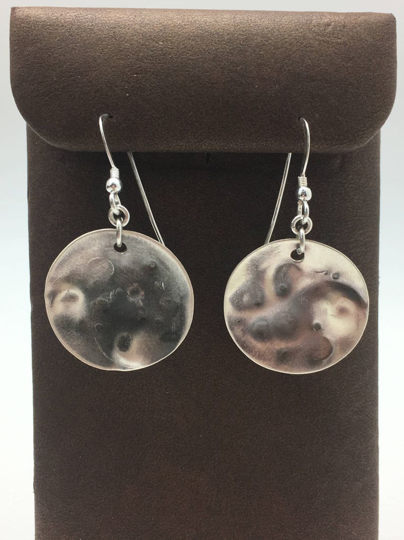 May include: A pair of silver dangle earrings with a textured, moon-shaped design. The earrings are hanging from a brown velvet background.