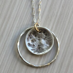 K&ouml;nnte beinhalten: Halskette mit silberfarbenem Anh&auml;nger. Der Anh&auml;nger hat ein strukturiertes, kreisf&ouml;rmiges Design, das an den Mond erinnert, und ist in einem geh&auml;mmerten Silberring aufgeh&auml;ngt. Die Kette hat eine feine Silberkette und eine goldfarbene &Ouml;se.