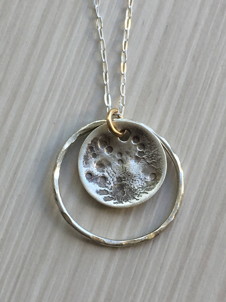 K&ouml;nnte beinhalten: Halskette mit silberfarbenem Anh&auml;nger. Der Anh&auml;nger hat ein strukturiertes, kreisf&ouml;rmiges Design, das an den Mond erinnert, und ist in einem geh&auml;mmerten Silberring aufgeh&auml;ngt. Die Kette hat eine feine Silberkette und eine goldfarbene &Ouml;se.