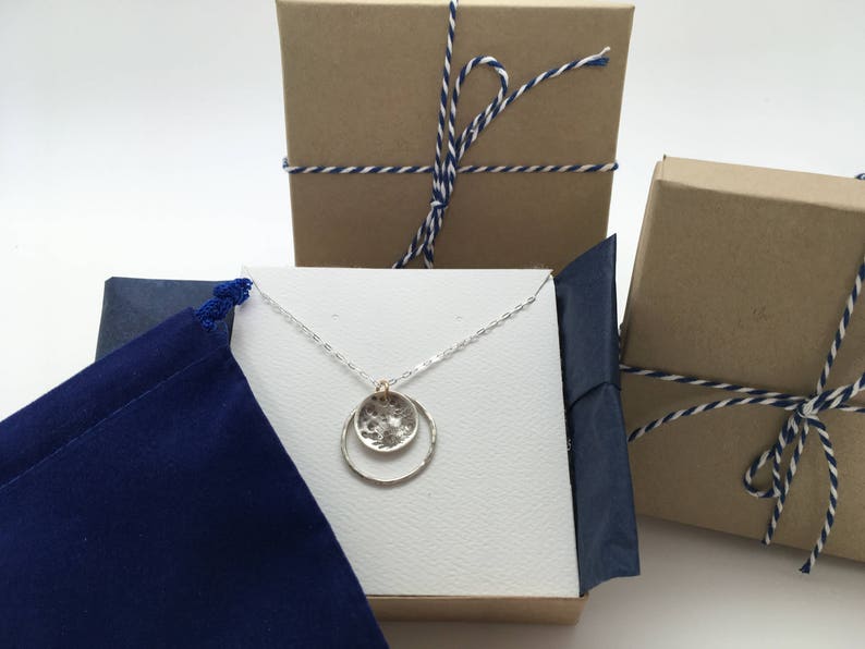 May include: A silver necklace with a textured circle pendant and a larger, smooth circle pendant. The necklace is displayed on a white background with a blue velvet pouch and brown boxes.