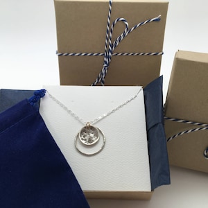 K&ouml;nnte beinhalten: Eine silberne Halskette mit zwei kreisf&ouml;rmigen Anh&auml;ngern, einer geh&auml;mmert und der andere glatt, pr&auml;sentiert in einer Geschenkbox. Die Box ist von zwei weiteren Geschenkboxen umgeben, die mit blau-wei&szlig;em Garn gebunden sind, und einem blauen Samtbeutel.