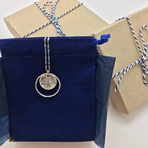 K&ouml;nnte beinhalten: Eine silberne Halskette mit einem geh&auml;mmerten Scheiben- und offenen Kreisanh&auml;nger h&auml;ngt an einem blauen Samtbeutel. Im Hintergrund befinden sich zwei verpackte Geschenke mit blau-wei&szlig;em Bindfaden.