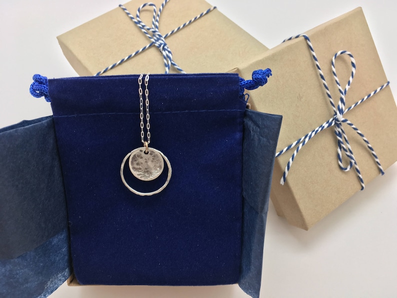 K&ouml;nnte beinhalten: Eine silberne Halskette mit einem geh&auml;mmerten Scheiben- und offenen Kreisanh&auml;nger h&auml;ngt an einem blauen Samtbeutel. Im Hintergrund befinden sich zwei verpackte Geschenke mit blau-wei&szlig;em Bindfaden.