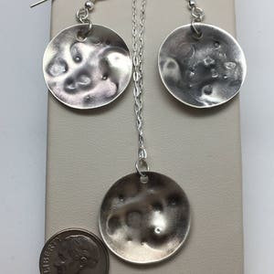 May include: A silver necklace and earrings set with round, textured pendants. The pendants are about the size of a US dime.